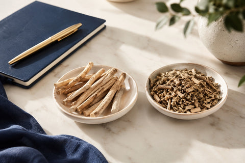 Two ceramic dishes of whole and ground ashwagandha root on marble beside a navy journal